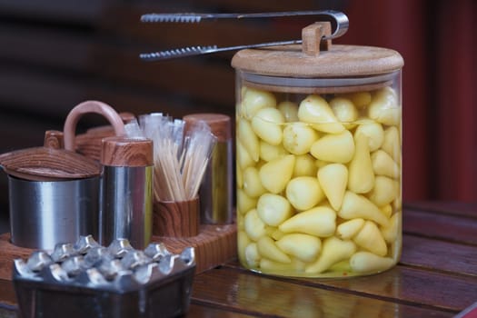 Yellow cloves of pickled garlic are displayed in a jar with utensils nearby.