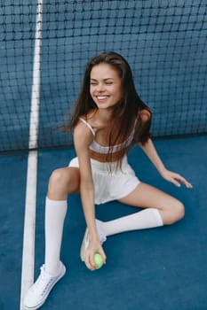 Cheerful young woman in sports attire sitting on a tennis court, expressing joy while holding a tennis ball, perfect for fitness and sports concepts