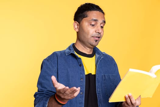 Indian male model reading a book to have fun and enjoy characters, standing with lecture against studio background. Casual guy browsing novel, enjoying leisure time and appreciating literature.