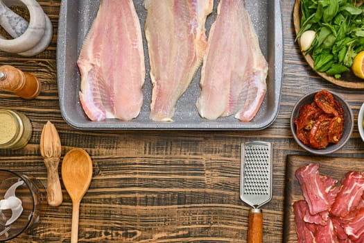 Fresh fish fillets are laid out on a baking tray, surrounded by various ingredients including herbs, lemon, and spices. Wooden utensils and bowls add a rustic touch to the kitchen workspace.