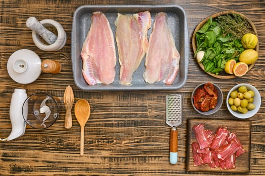 Various fresh ingredients are arranged on a wooden tabletop, including fish fillets, herbs, olives, and spices. This setup is perfect for creating a delightful seafood meal.
