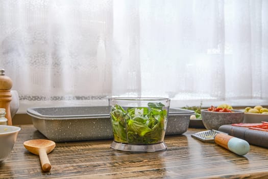 Fresh greens are placed in a chopping bowl of immersion blender on a wooden countertop alongside various ingredients. Sunlight filters through the curtains, enhancing the cozy kitchen atmosphere.