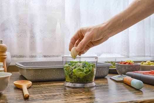 A person is adding a clove of garlic to a bowl filled with fresh greens, highlighting the process of meal preparation. The cozy kitchen features wooden surfaces and natural light streaming in.