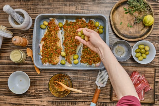 A hand adds green olives to a tray of seasoned fish fillets covered in vibrant herbs and spices. Surrounding the dish are various ingredients, creating a warm cooking atmosphere.