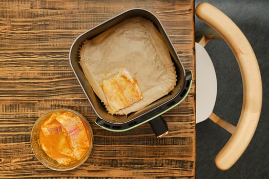 Sliced fish seasoned with spices sits on parchment paper inside an air fryer, ready for cooking. A glass bowl nearby contains additional fish pieces marinated in spices, all set on a wooden table.