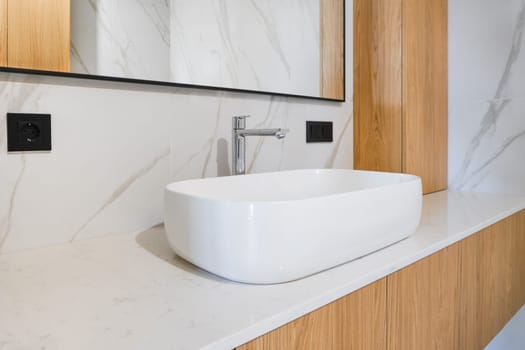 A contemporary bathroom showcases a sleek, white oval sink placed on a light marble countertop. Natural wood elements complement the elegant mirror and minimalist fixtures in this stylish space.