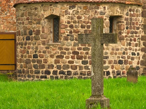 A historic stone cross in front of an old rustic church with small windows. Lively green grass in the foreground adds depth and contrast to the picture.