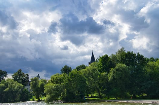 Green river with lush trees, dramatic sky and church tower in the background. Harmonious landscape, ideal for themes such as nature and architecture.