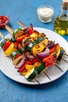 Grilled vegetable skewers featuring bell peppers, zucchini, onions, and mushrooms arranged on a decorative plate. Accompanied by sauces for dipping and olive oil in a glass bottle.