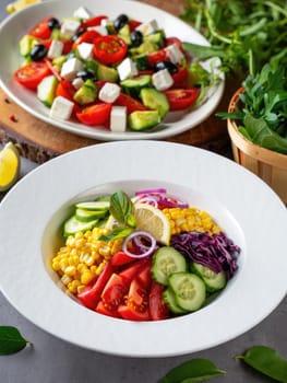 A vibrant salad bowl filled with corn, cucumber, tomatoes, red cabbage, and lemon slices sits on a dining table. In the background, a second salad featuring diced vegetables is visible.