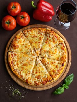 Freshly baked cheese pizza is sliced and served on a wooden board alongside ripe tomatoes, a red pepper, and a glass of red wine, creating a charming culinary display.