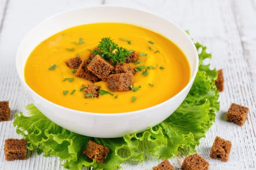 A vibrant bowl of creamy pumpkin soup topped with crunchy croutons and garnished with parsley sits on a bed of fresh lettuce. This appetizing dish is ideal for a cozy meal.