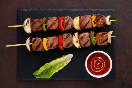Juicy grilled beef skewers are arranged carefully with bell peppers, onions, and a fresh lettuce leaf on a dark slate platter. A small bowl of tangy dipping sauce complements the dish.
