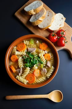 A warm bowl of pasta soup features spiral noodles, carrots, and tender meat, garnished with fresh herbs. Slices of bread and tomatoes accompany the dish on a wooden cutting board.