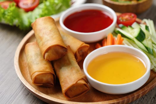 A wooden plate presents multiple golden brown spring rolls accompanied by two dipping sauces—sweet and spicy—alongside fresh vegetables like cucumbers, carrots, and lettuce.