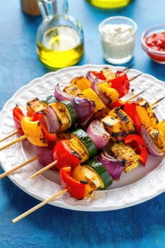 Grilled vegetable skewers showcase a mix of bell peppers, zucchini, and onions on wooden sticks. They are served on a white plate with olive oil, dips, and a vibrant blue background.