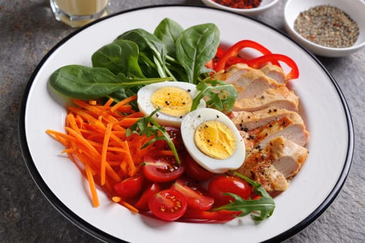 A colorful salad features fresh spinach, sliced carrots, cherry tomatoes, and grilled chicken breast served with boiled eggs. A variety of spices accompanies the dish, enhancing its appeal.