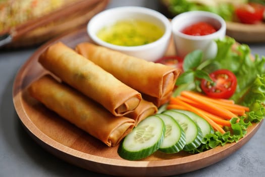 Golden spring rolls are plated alongside fresh cucumber slices, lettuce, tomatoes, and carrots. Two bowls contain dipping sauces, enhancing the flavorful presentation.