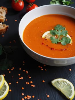 A bowl of rich red lentil soup is presented with lemon slices and fresh cilantro on top. Surrounding the bowl are scattered lentils, fresh tomatoes, and slices of bread, creating an inviting meal.