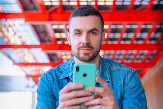 Confident man in denim jacket holding smartphone and looking directly at the camera. Portrait in modern urban setting with red industrial beams and strong daylight. Stylish and focused expression.