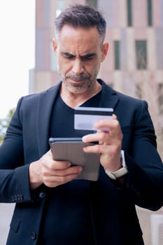 Confident caucasian man with headphones serious while using tablet and holding a credit card outdoors. Concept of online shopping, digital payment, modern lifestyle, and financial technology. Vertical