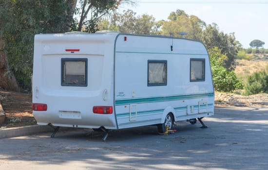 A white caravan rests beside a tranquil dirt road flanked by greenery and trees. The sunlight casts a warm glow, inviting exploration and relaxation in this peaceful setting.