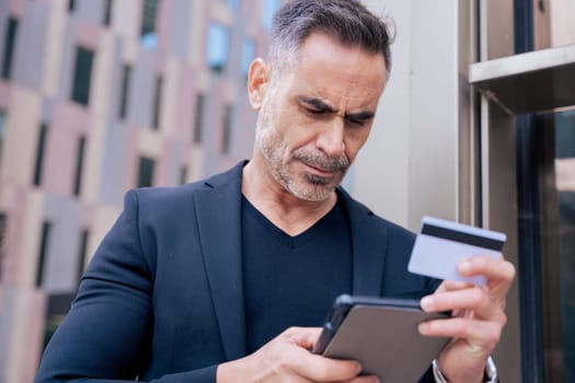 Confident caucasian man with headphones serious while using tablet and holding a credit card outdoors. Concept of online shopping, digital payment, modern lifestyle, and financial technology.