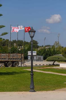 Vibrant green grass meets classic street lamppost, adorned by Turkish and northern Cyprus flags fluttering gently against a clear blue backdrop, inviting leisurely strolls.
