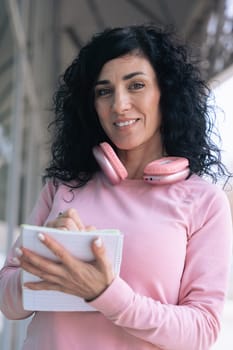 Athletic woman with curly black hair, wearing a pink hoodie and headphones, looking at the camera while writing in a notebook. She stands outdoors in an urban setting, confident,focused, and motivated