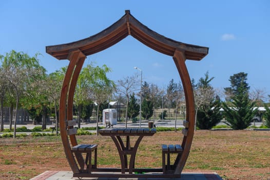 A wooden gazebo stands invitingly in a sunny park, surrounded by lush greenery and gentle trees. The clear blue sky enhances the serenity, making it a perfect place to unwind.