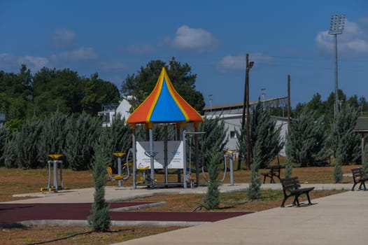 A colorful exercise station stands prominently in a peaceful park, surrounded by lush trees and scattered benches, inviting visitors to engage in fitness activities under bright sunlight.