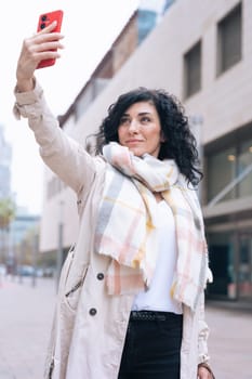 Confident woman wearing scarf and light jacket takes a selfie with red smartphone on a city street. Modern urban buildings in the background. Expression of self-assurance and personal style. Vertical