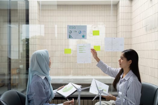 In a glass-walled office, two women engage in a dynamic discussion about ESG initiatives, analyzing data and sharing insights to enhance sustainable practices.