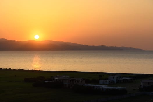 Vibrant hues of orange and yellow fill the sky as the sun dips below tranquil waters. Distant mountains create a serene backdrop, with silhouettes of houses surrounded by lush greenery, evoking peace.