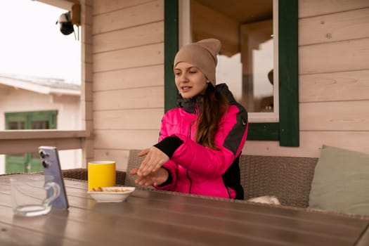 A young woman wearing a vibrant jacket sits on a porch, sharing a snack while engaging in conversation with someone on her phone. The setting features a wooden cabin and cloudy skies.
