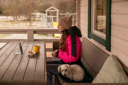 A young woman wearing a bright pink jacket sits quietly on a porch, sipping coffee while her dog lounges beside her. The peaceful countryside setting adds to the serene atmosphere.