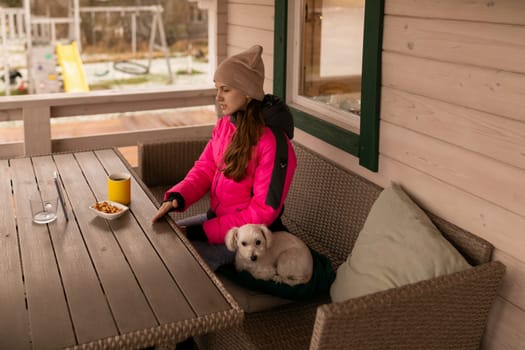 A young woman wearing warm clothing sits on a porch with her small dog beside her. She is relaxing on a cushioned bench, sipping a drink and surrounded by nature.
