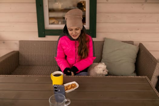 A woman in a pink jacket sits joyfully at a table on a winter afternoon, enjoying a cup of coffee and a plate of snacks while her small dog cuddles beside her.