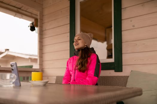 A woman wearing a pink jacket and a beanie sits comfortably at a wooden cabin, enjoying a warm drink as she takes in the tranquil outdoor atmosphere on a chilly day.