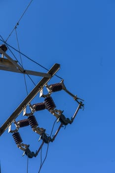 High above, a power distribution system supports cables against a bright blue sky. Metal components glisten under sunlight, showcasing modern engineering in an urban setting.