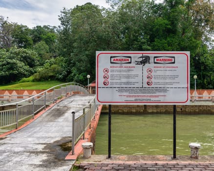 A warning sign advises about the presence of crocodiles beside a tributary of Sungai Brunei, or Brunei River in Bandar Seri Begawan. The area features a bridge and lush greenery