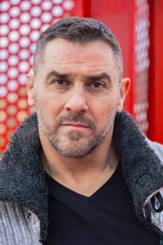 Portrait of a serious caucasian man standing by a bold red metal structure in an urban environment. Wearing a gray sweater and black shirt, he looks confidently at the camera with city view behind