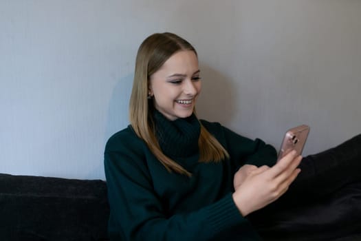 A young woman with long hair is sitting on a couch, smiling as she interacts with her smartphone. The cozy indoor environment suggests a relaxed moment.