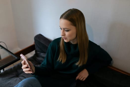 A young woman with long hair is sitting comfortably on a couch, engaged with her smartphone. Soft daylight illuminates her focused expression in a minimalistic indoor space.