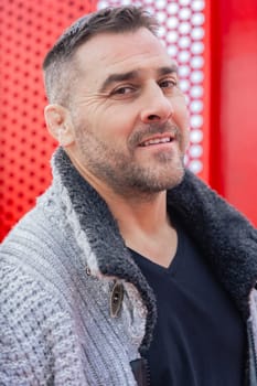 Close-up individual portrait of a middle-aged man with short hair and beard, wearing a winter outfit. Smiling expression, urban background with red metal structure, conveying confidence and depth.