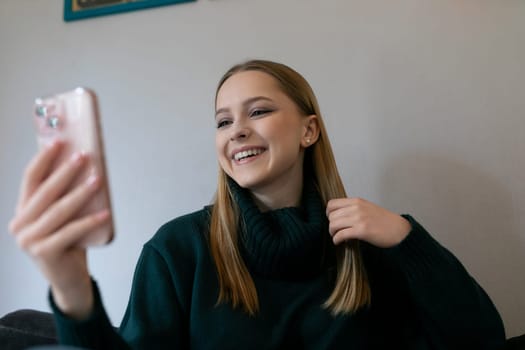 A young woman sits comfortably on a couch, smiling as she engages in a virtual conversation on her smartphone. The cozy living room features soft lighting and neutral colors.