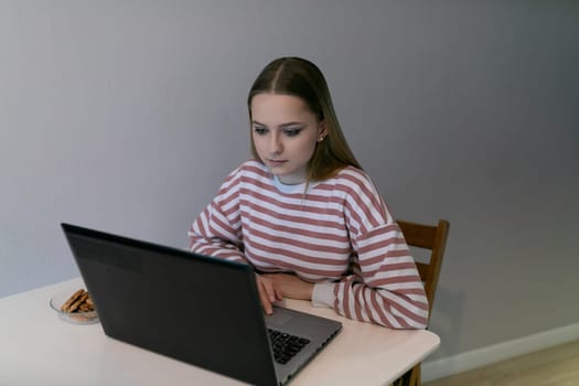 Focused young woman engages with her laptop, immersed in work or study, while seated at a table with cookies nearby in a cozy, minimalist room.