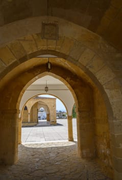 Stone arches create a beautiful passageway, leading to a tranquil courtyard. Bright sunlight illuminates the intricate architecture, inviting wanderers to explore its history.