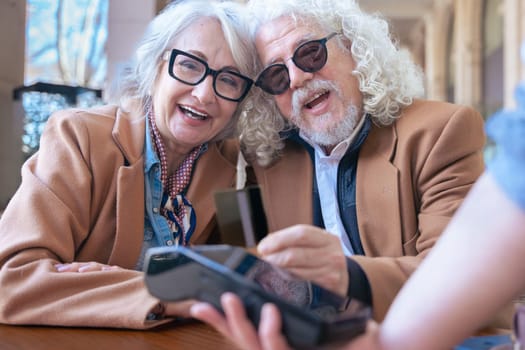 Joyful caucasian senior couple at an outdoor cafe making a card payment. Both wearing trendy outfits and sunglasses. Ideal for retirement, finance, lifestyle, and senior couple relationship concepts.