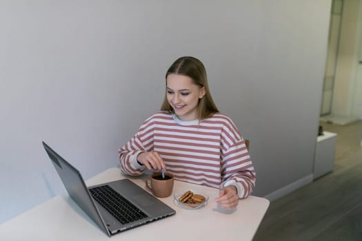 A woman in a striped sweater smiles while dipping a cookie into her hot drink at a small table. She is focused on her laptop in a light and airy room.